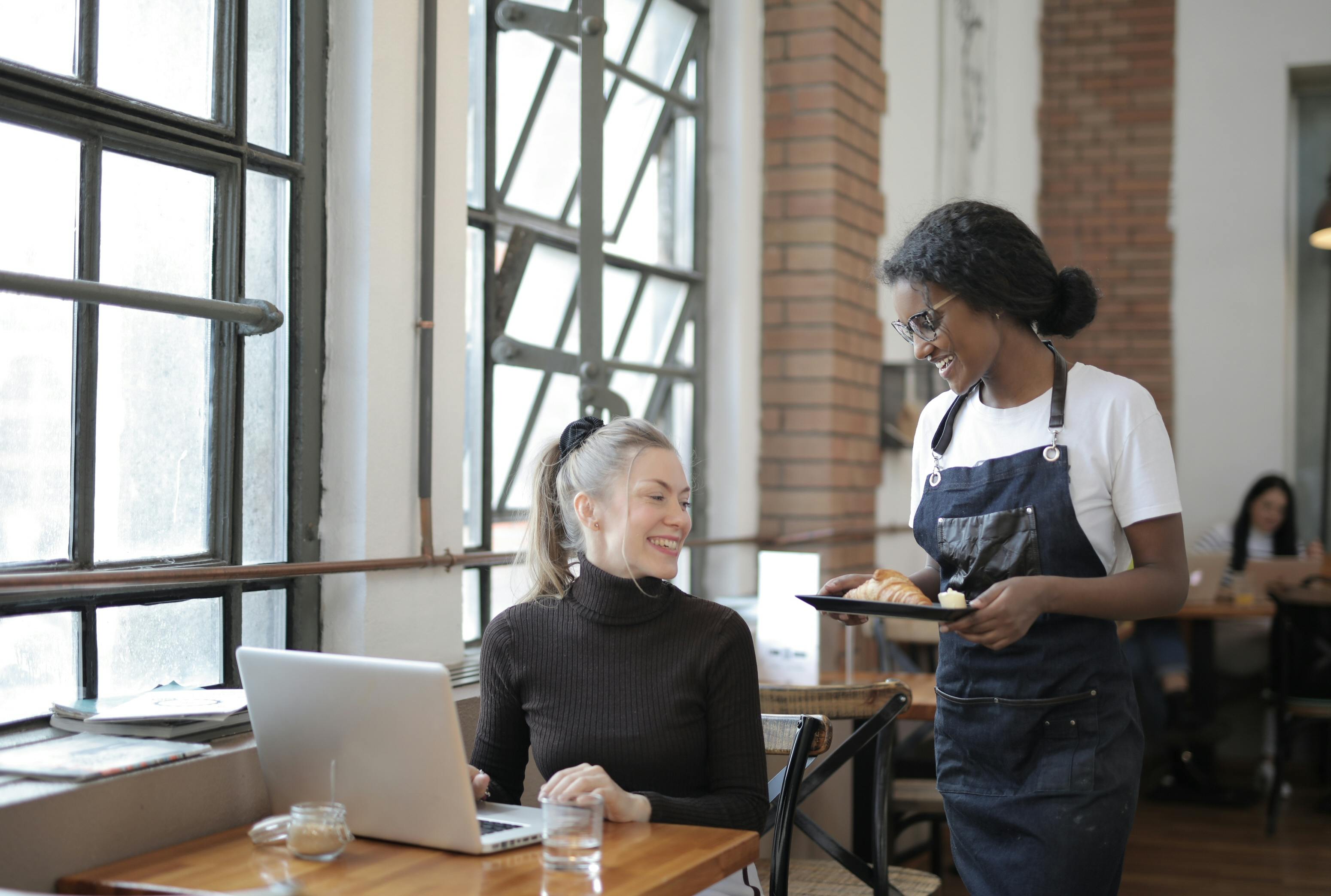 Two women in a casual office setting with one using a laptop and the other holding a tray.